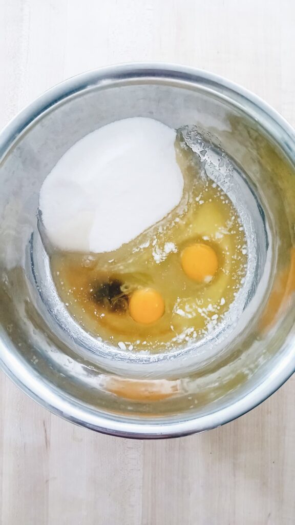 Apple cake wet ingredients. A metal mixing bowl contains two raw eggs, sugar, and vanilla extract on a wooden surface.