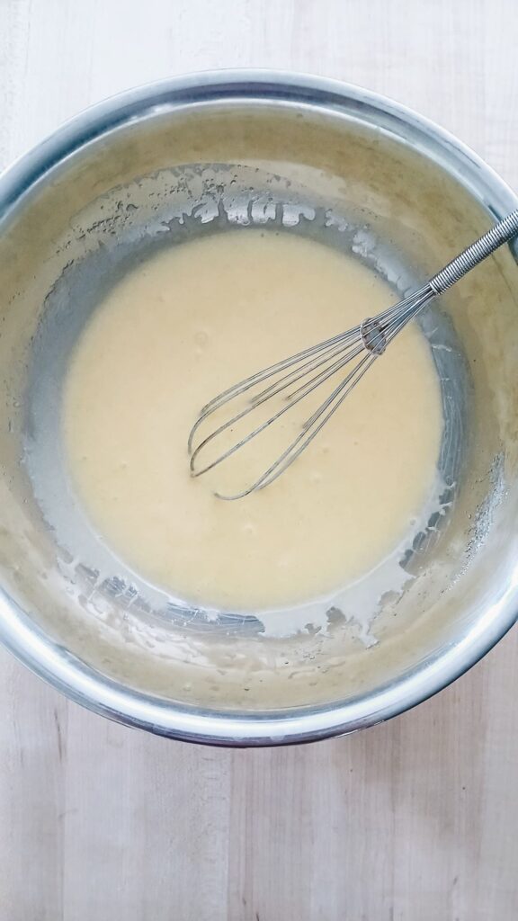 A metal whisk rests in a stainless steel bowl filled with smooth, creamy apple cake wet ingredients that have been mixed together.
