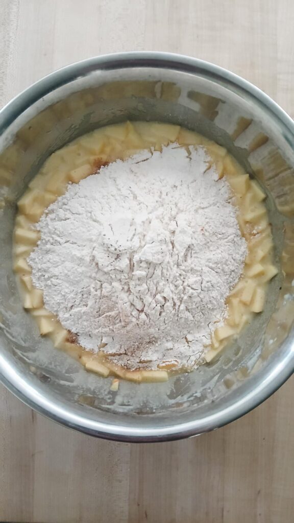 A metal mixing bowl filled with chopped apples, covered by a pile of flour. Adding the dry ingredients to the wet ingredients when making apple cake and ready to mix