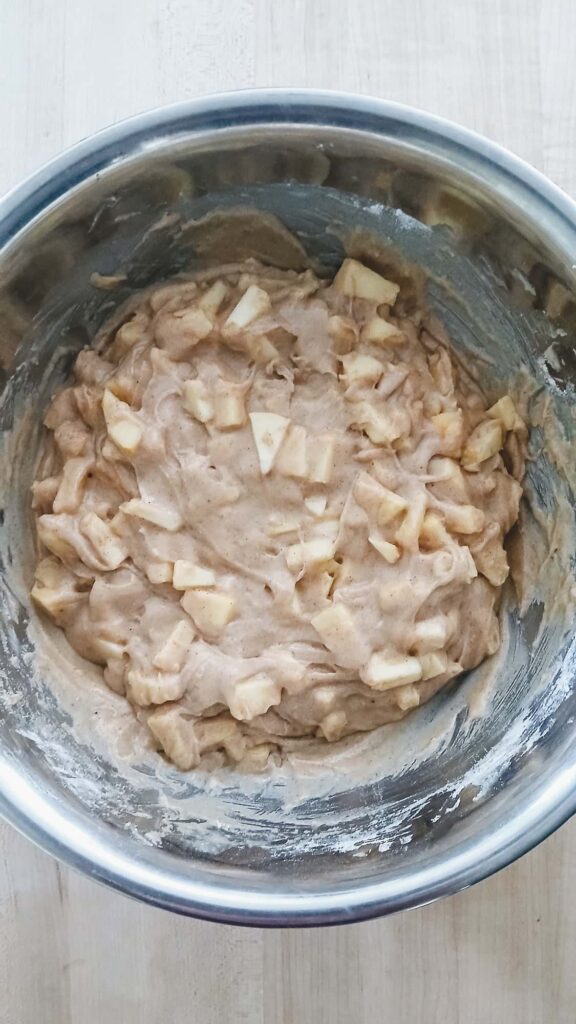All the ingredients for apple cake batter mixed together in a metal bowl. The batter is cinnamon color with visible apple chunks.