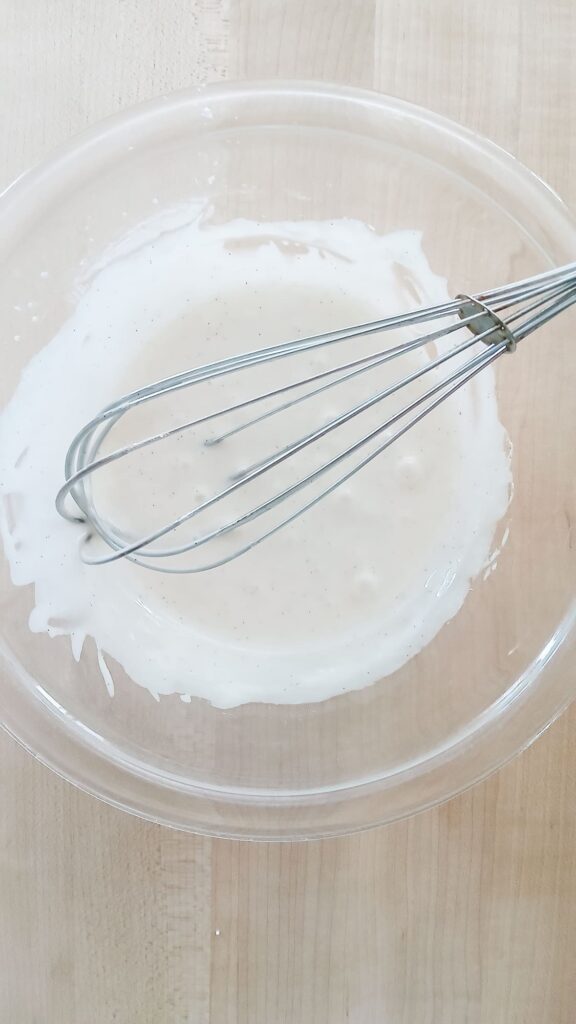 A metal whisk rests in a glass bowl, stirring a smooth powdered sugar glaze that will go on top of apple cake