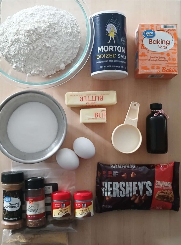 A top-down view of all ingredients needed for chai sugar cookies: bowls of flour, granulated sugar, butter, two eggs, various chai spices, and cinnamon chips, neatly arranged on a kitchen counter.