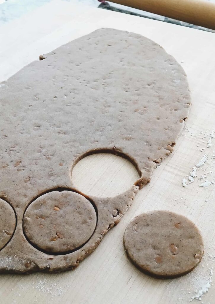 A sheet of chai sugar cookie dough on a lightly floured surface. Several round cookies have been cut out, with a wooden rolling pin visible in the background.