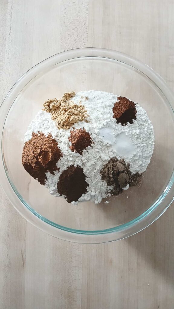 A clear glass mixing bowl containing flour. On the surface of the flour are distinct, visible mounds of each chai spice, including cinnamon, cardamom, ginger, cloves, and allspice, before being mixed.