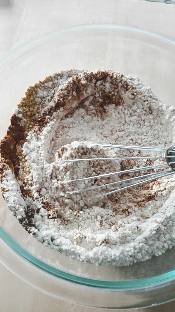 A whisk is partially submerged in a clear bowl of dry ingredients, blending the chai spices directly into the flour for the spiced sugar cookie dough.