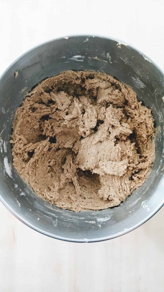 A top-down view of the unbaked cookie dough in a metal stand mixer bowl. The dough is thick and has a rich, medium-brown color due to the ground chai spices being thoroughly mixed in.