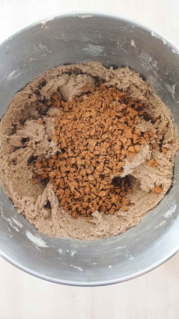 The spiced, brown cookie dough in a silver stand mixer bowl, topped with a pile of chopped cinnamon chips ready to be folded in.