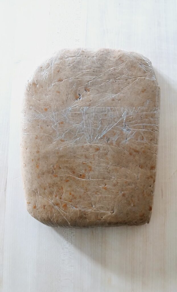 The finished chai sugar cookie dough, shaped into a flat square and tightly wrapped in clear plastic wrap, ready to be placed in the refrigerator to chill.