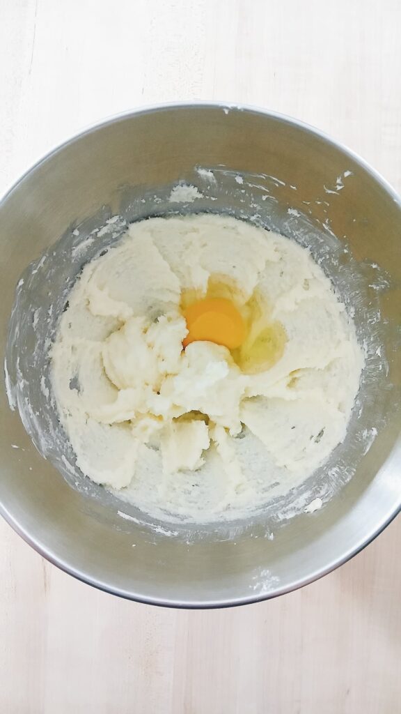 An egg is cracked into the center of the creamed butter and sugar mixture in a metal stand mixer bowl, just before it is mixed in for the cookie dough.