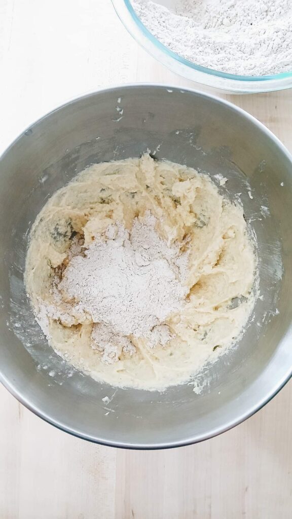 A top-down view of a metal stand mixer bowl containing the creamed butter and egg mixture, with the whisked flour and chai spice blend added on top, ready to be mixed