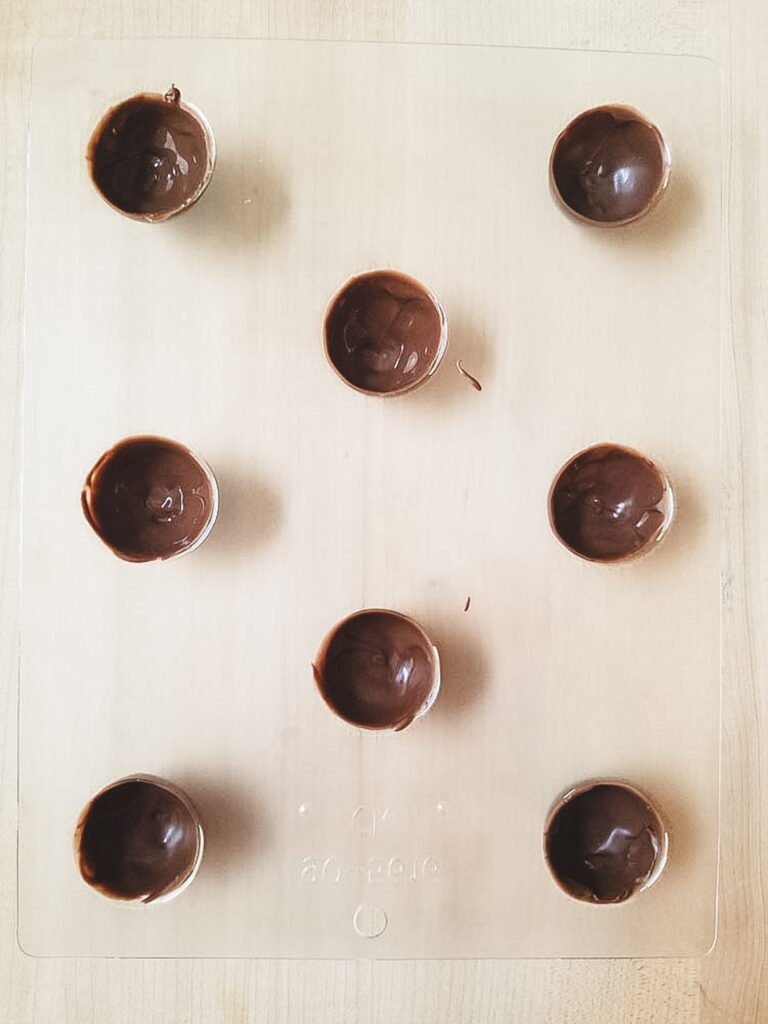 A clear mold with eight small, round cavities each partially filled with melted chocolate. This is the mold to make the chocolate shell for chocolate covered cherries.