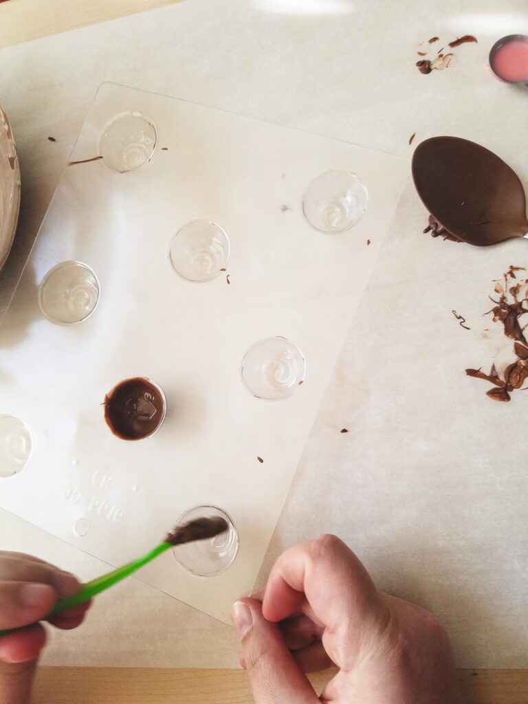 Hands are seen using a baby spoon to coat the inside of a candy mold with chocolate in order to make a candy shell for chocolate covered cherries