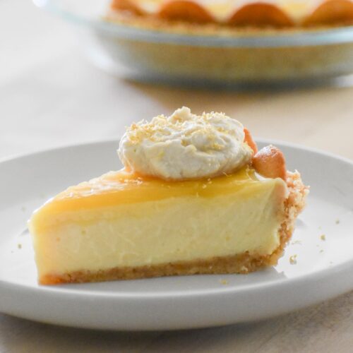 A slice of creamy lemon icebox pie on a white plate, topped with whipped cream and lemon zest. The pie has a crumbly crust, with a whole pie in the background.