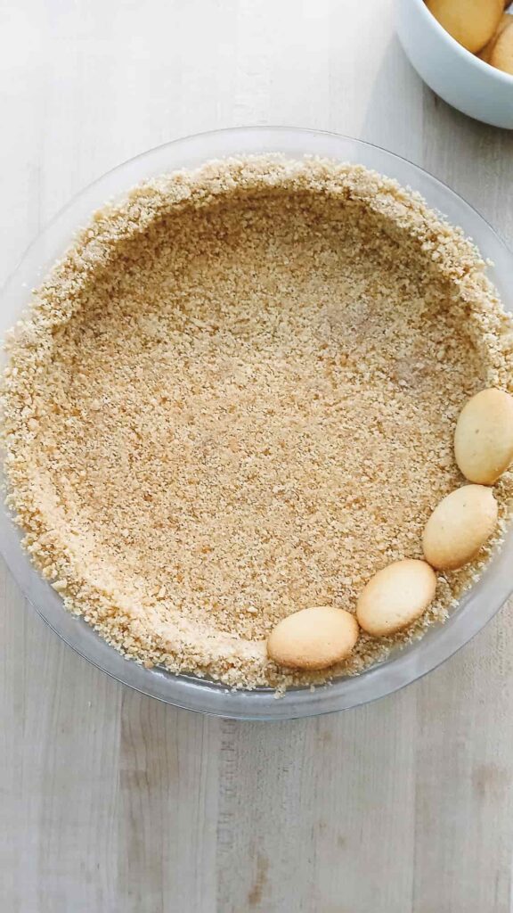 A clear glass pie dish filled with fine vanilla wafer crust sits on a light wooden surface. Three vanilla wafers rest on the crust's edge starting to make the decorative border of the crust