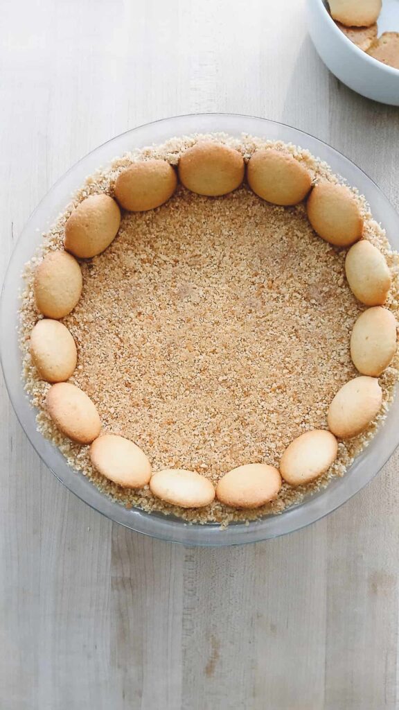 A pie crust topped with a ring of vanilla wafer cookies and filled with finely crushed crumbs, set on a light wooden surface. A bowl of similar cookies is in the background.