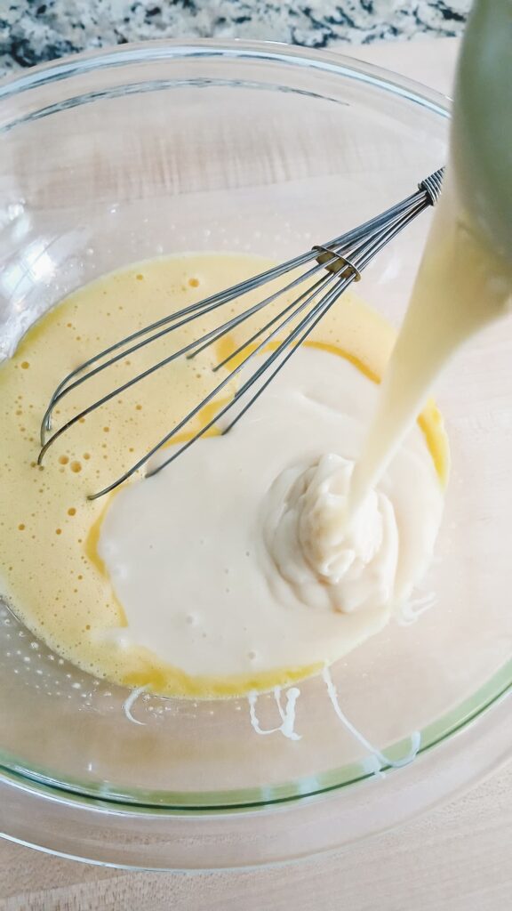 Whisk in a glass bowl with beaten eggs and condensed milk is being poured in