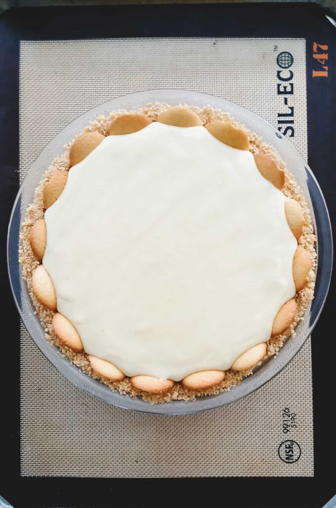 A round lemon icebox pie with a smooth creamy top, bordered by a layer of vanilla cookies, sits on a baking mat. Ready to go into the oven to be baked