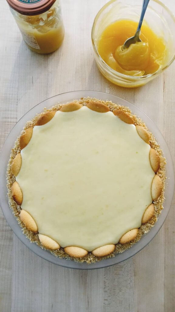 A chilled lemon icebox pie ready to be topped. Nearby are a jar and bowl of lemon curd.