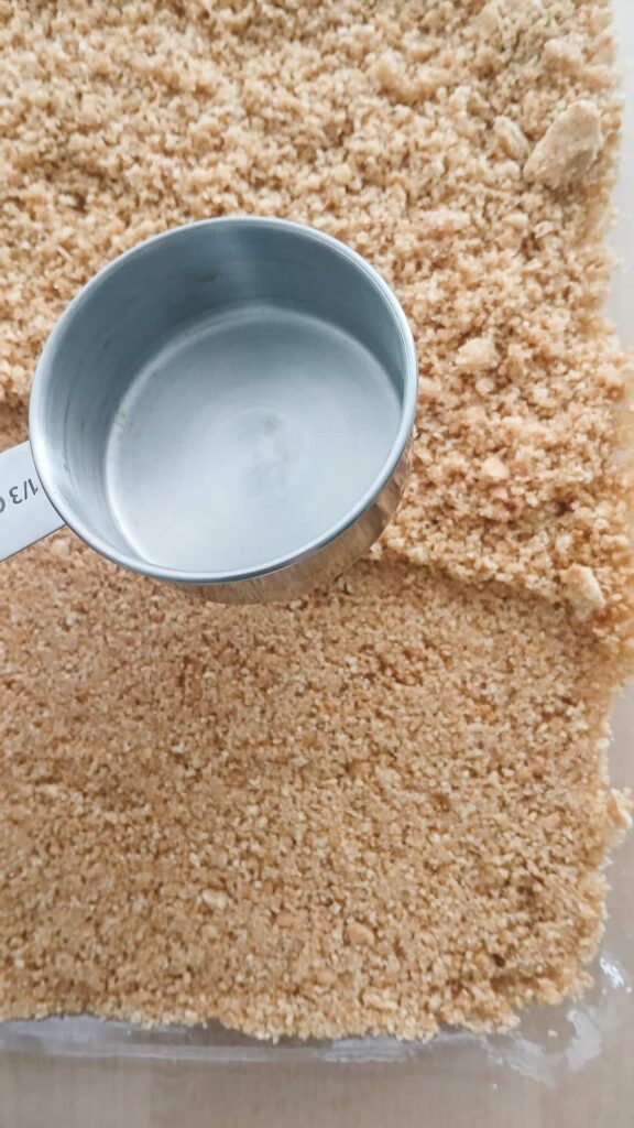 Close up of a metallic measuring cup resting on finely crushed graham crackers. The texture is coarse, with some larger chunks visible.