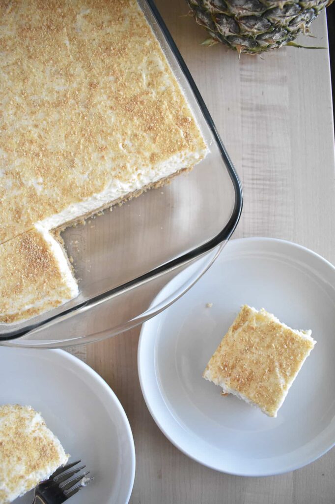 Overhead view of pineapple dessert in a glass dish, with one piece served on a white plate. Nearby, a whole pineapple rests on a light wooden table.