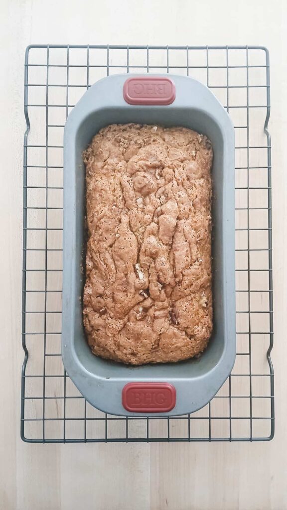 Golden brown pistachio bread loaf resting in the pan on a wire cooling rack, showing the crunchy, toasted pecan and cinnamon sugar crust on top.