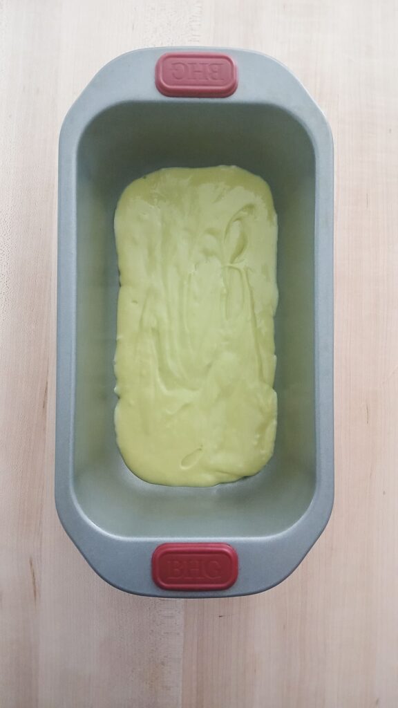 An overhead shot of a prepared nonstick 9x5 loaf pan with the first thick layer of pale green pistachio bread batter spread evenly across the bottom.