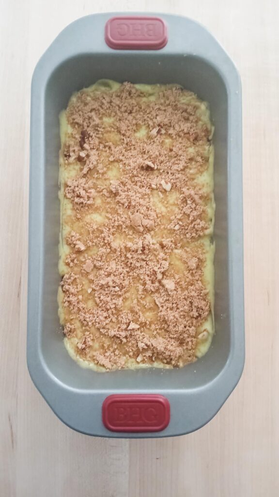 The final layer of thick pistachio batter is topped with a rustic crust of brown sugar and pecan crumble, showing the loaf prepped and ready for the butter knife swirling technique.