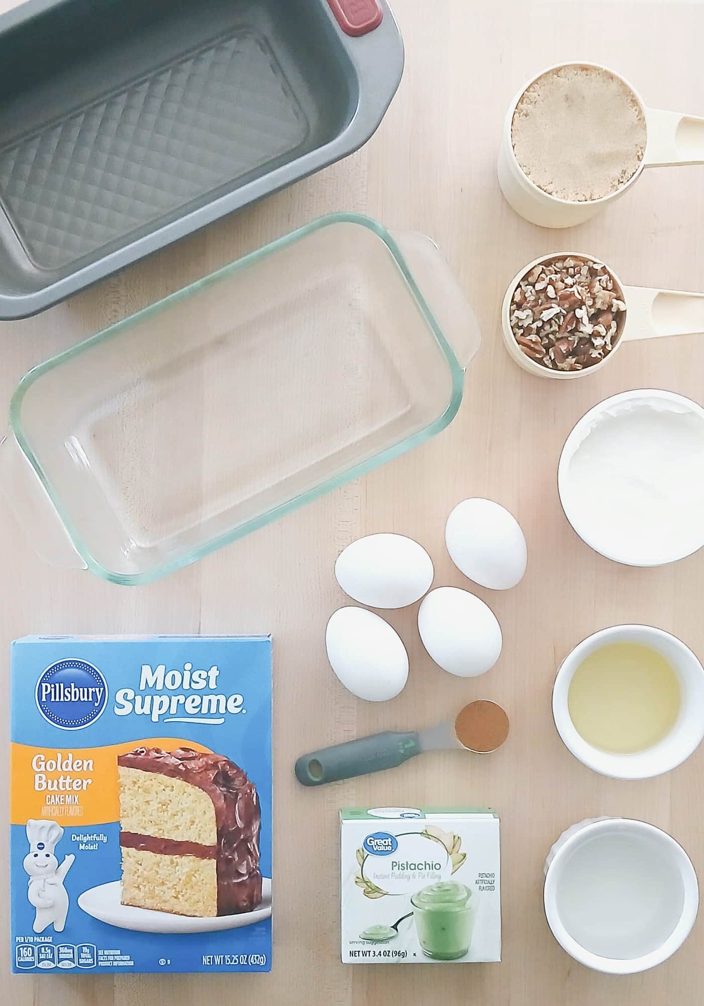Baking ingredients on a wooden surface include a cake mix box, pistachio pudding, three eggs, brown sugar, pecans, oil, and cake pans.