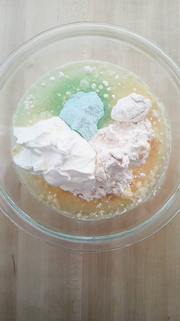 A overhead view of a mixing bowl containing golden butter cake mix, a pale green pistachio pudding mix, and a well of vegetable oil and water ready to be combined.