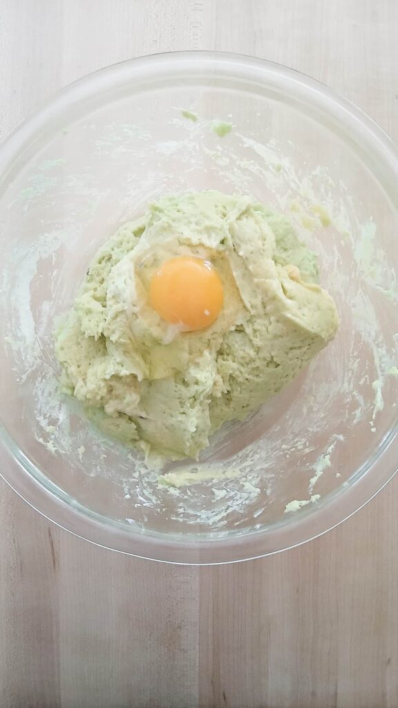 A mixing bowl with a single large egg added to the thick green pistachio batter to show the gradual emulsification process.