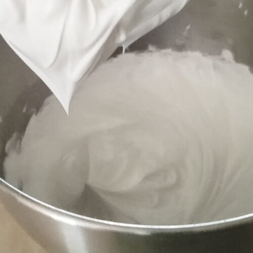 Royal icing forming stiff peaks in a metal mixing bowl, with a mixer whisk attachment above.