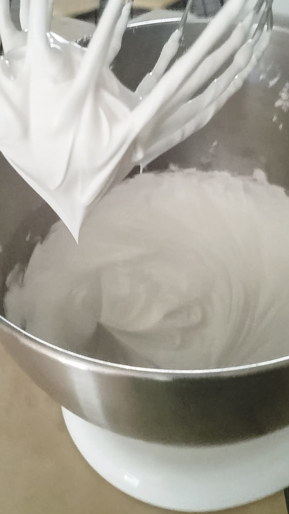 Royal icing forming stiff peaks in a metal mixing bowl, with a mixer whisk attachment above.