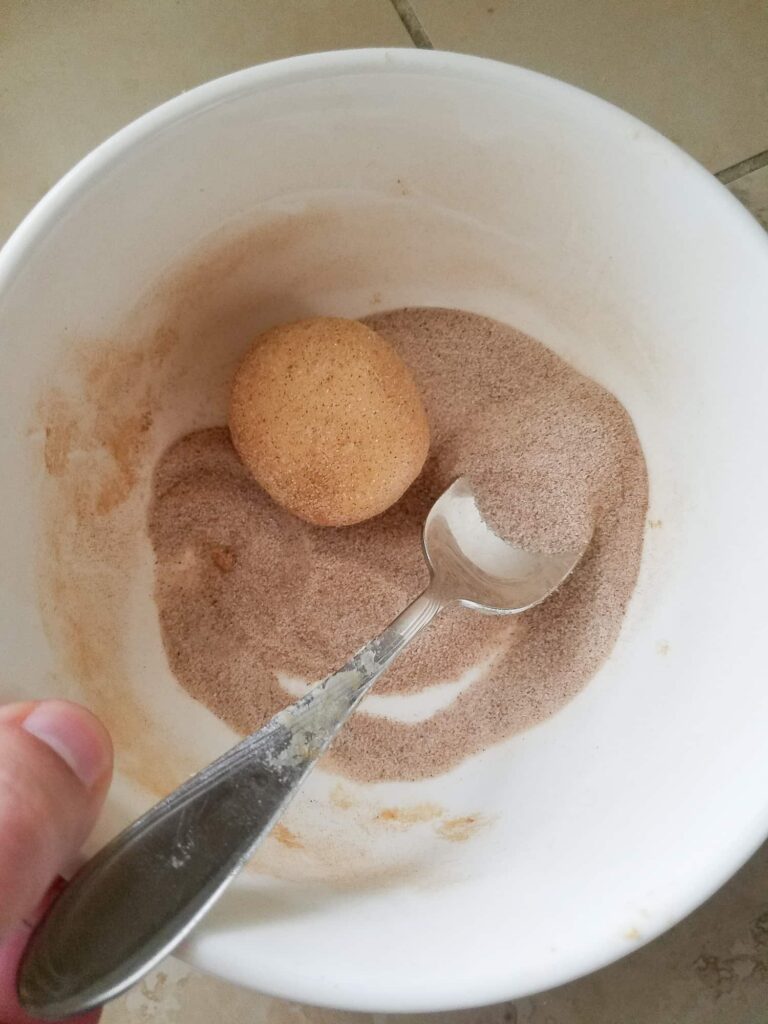 Using a spoon to toss a large snickerdoodle dough ball in a small bowl of cinnamon sugar coating for even coverage.