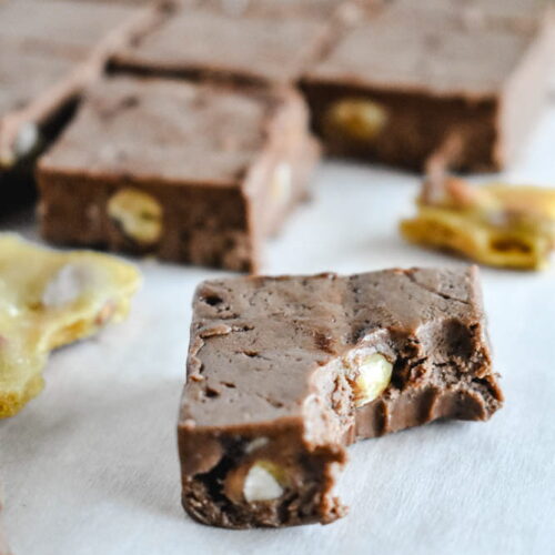 A close-up of a smooth chocolate fudge square with a bite taken out, showing the thick, creamy interior and crunchy peanut brittle pieces.