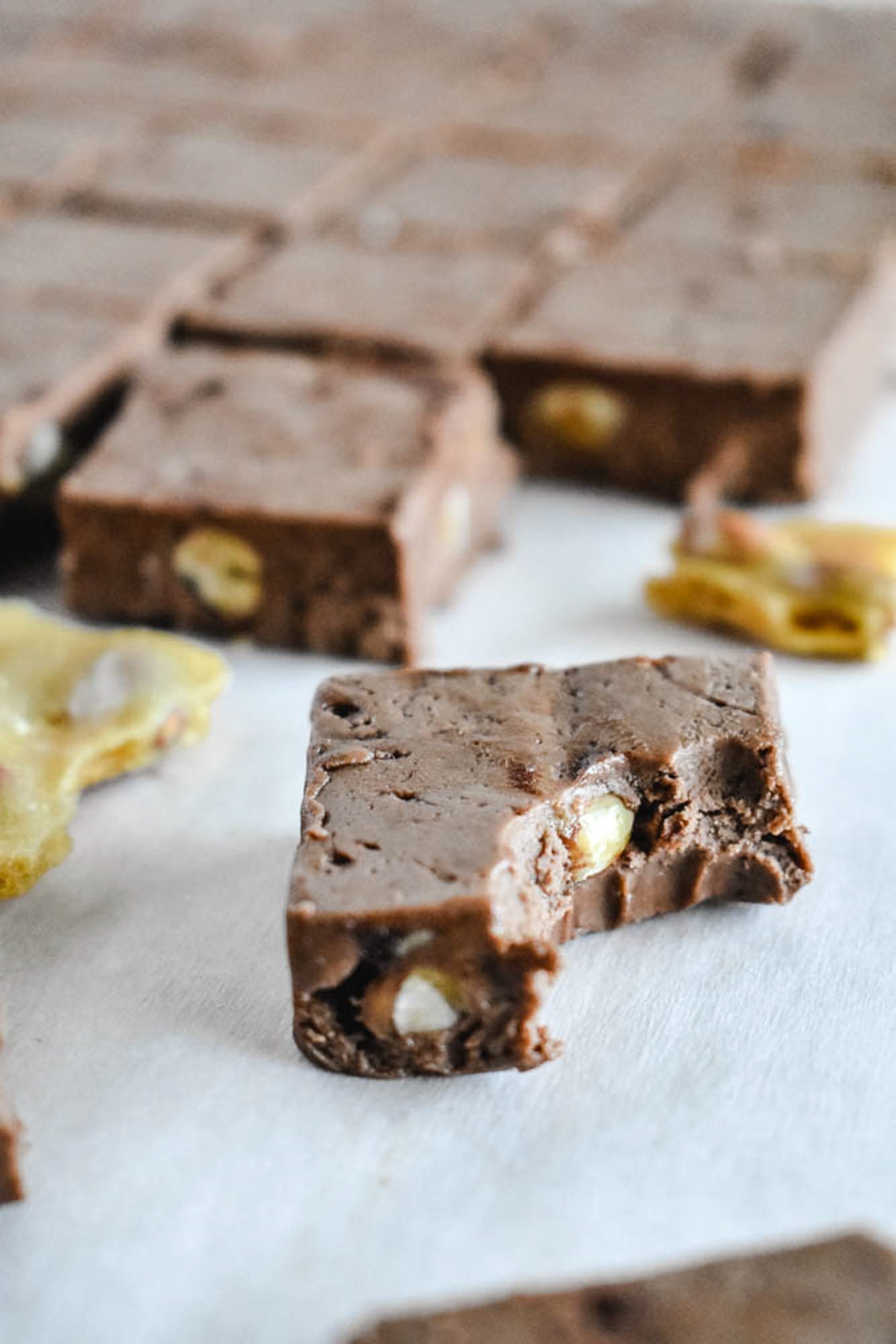 A close-up of a smooth chocolate fudge square with a bite taken out, showing the thick, creamy interior and crunchy peanut brittle pieces.
