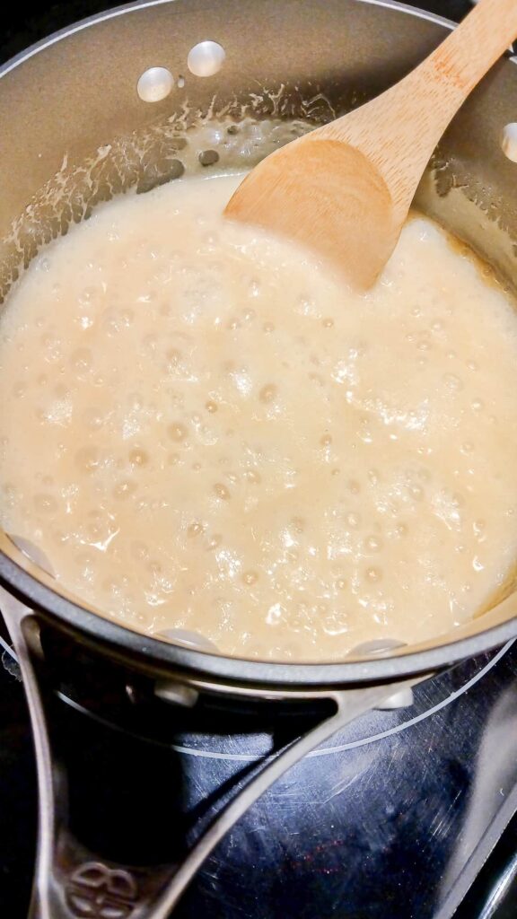 A heavy saucepan on the stove showing a rolling boil with thick, deliberate bubbles as the sugar mixture reaches the soft-ball stage for fudge