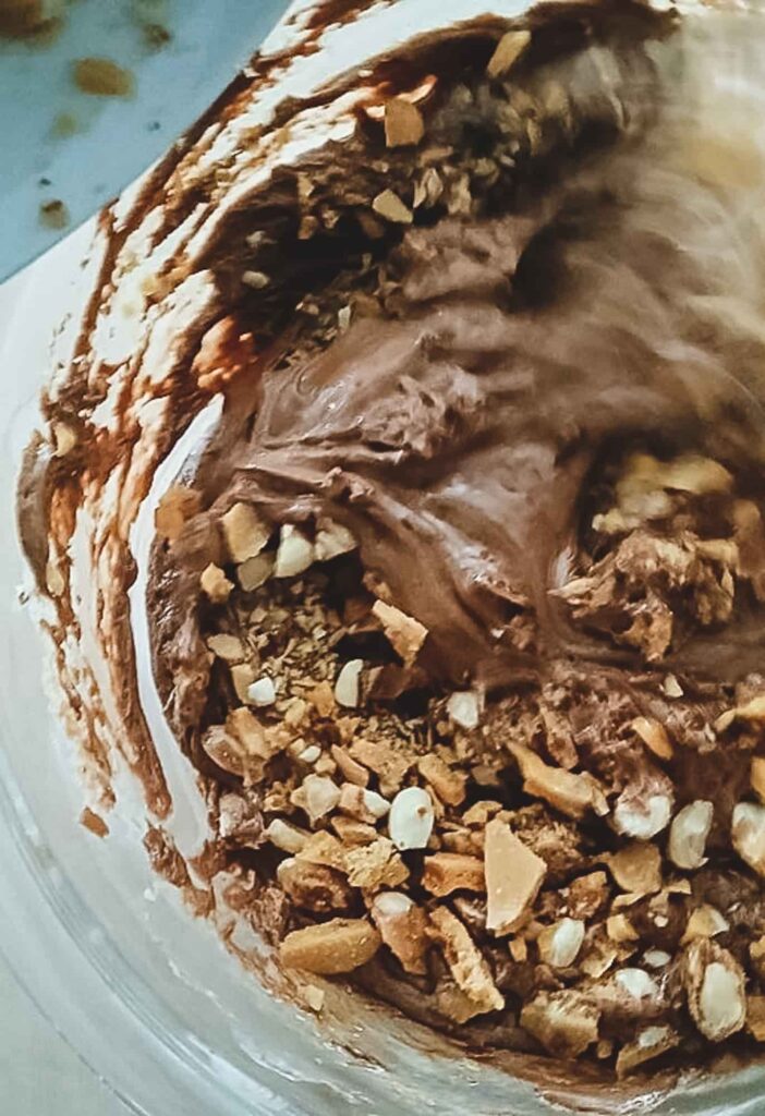 A close up of chopped peanut brittle shards sitting on top of the glossy chocolate mixture, ready to be folded in for a crunchy chocolate fudge