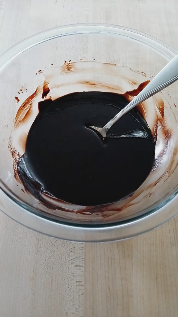 A dark, glossy chocolate mixture in a glass bowl with a metal spoon, showing the nearly black color of the melted butter and cocoa powder.