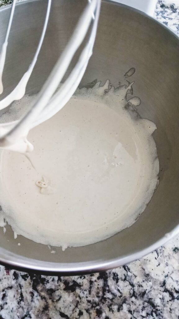 The egg and sugar mixture in a stand mixer after a six-minute whip, now a very pale cream color and thick, frothy texture similar to meringue.