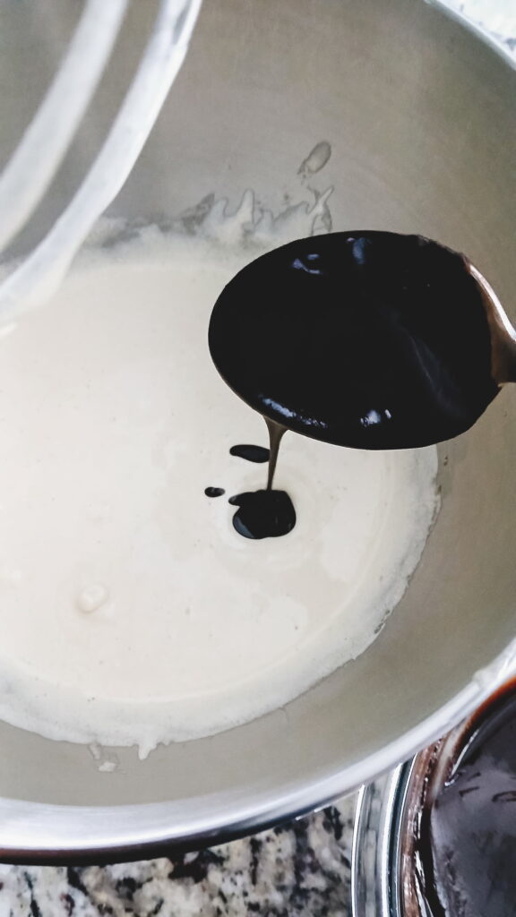 A metal spoon pouring the dark, glossy chocolate mixture into the pale and frothy whipped egg base in a stand mixer bowl.