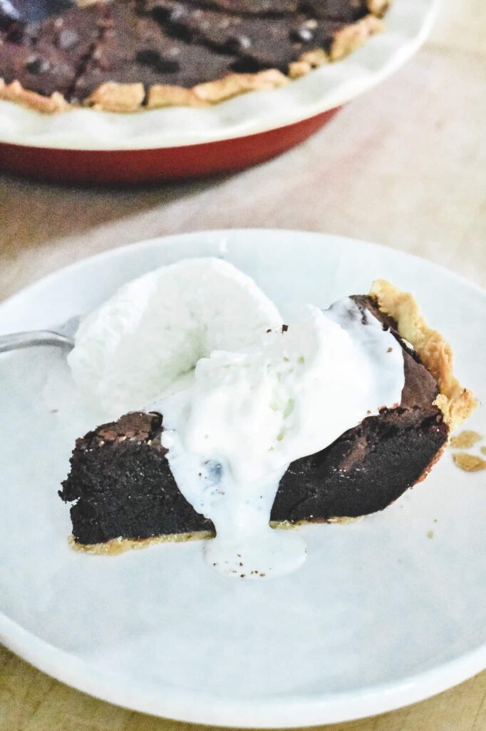 A slice of fudgy chocolate brownie pie on a white plate, topped with a scoop of melting vanilla ice cream, with the rest of the pie visible in the background.