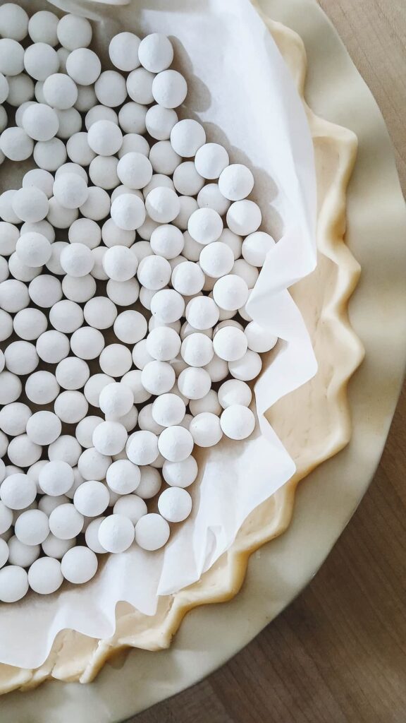 Close up of a raw, crimped pie crust lined with a circle of parchment paper and filled to the edges with white ceramic pie weights.