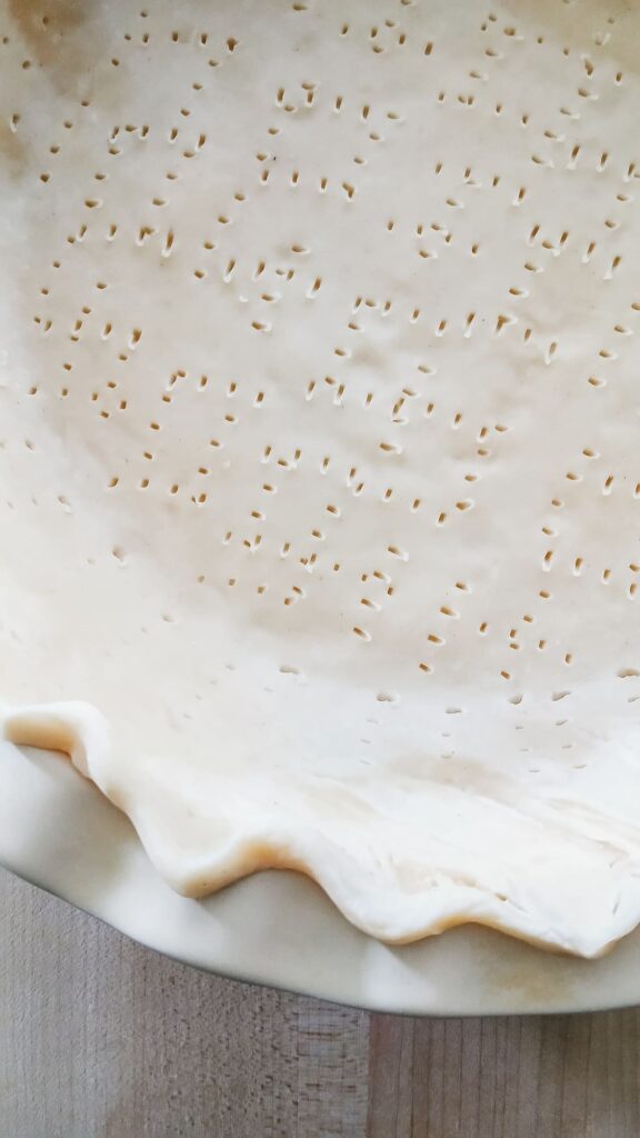 A 9-inch pie crust in a pan with the bottom and sides poked with fork marks to dock the dough before blind baking.