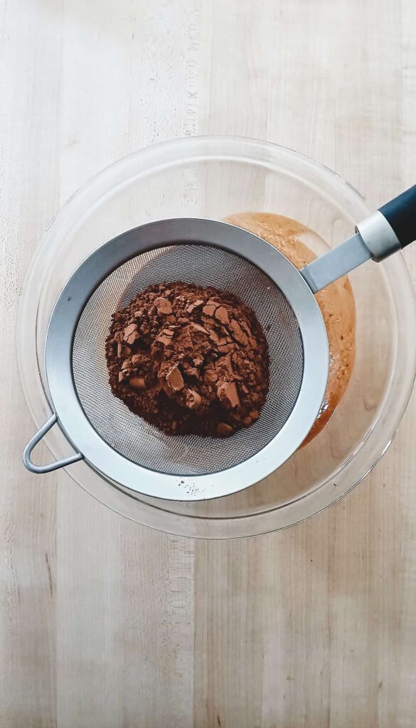 A fine-mesh sieve filled with dark cocoa powder held over a bowl of melted butter and chocolate.