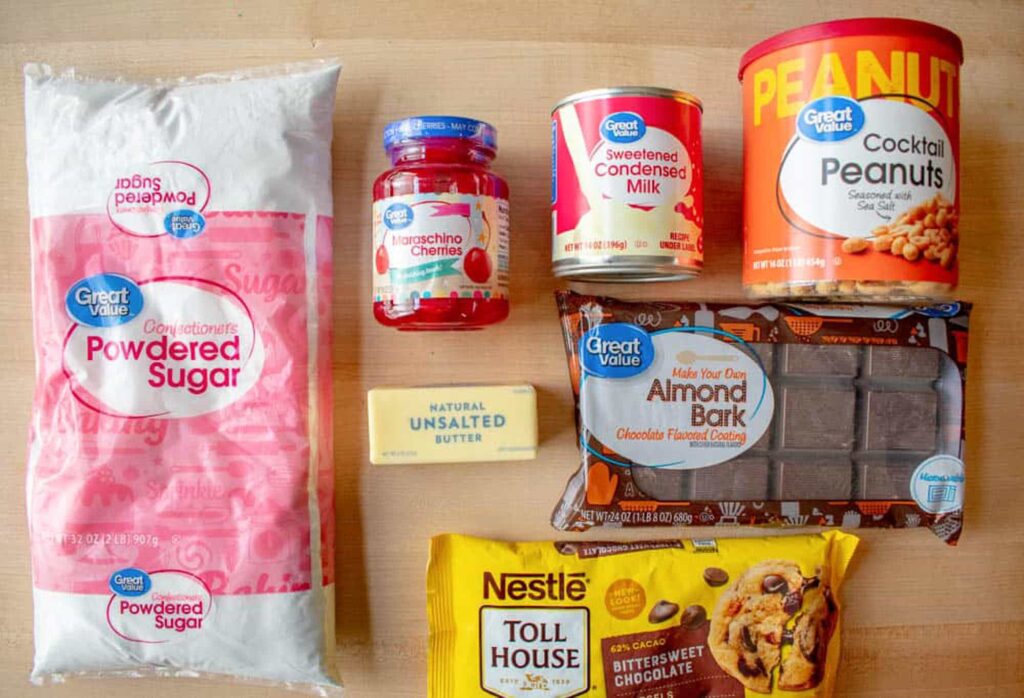 Candy making ingredients on a wooden surface: powdered sugar, maraschino cherries, unsalted butter, condensed milk, cocktail peanuts, almond bark, and chocolate chips.