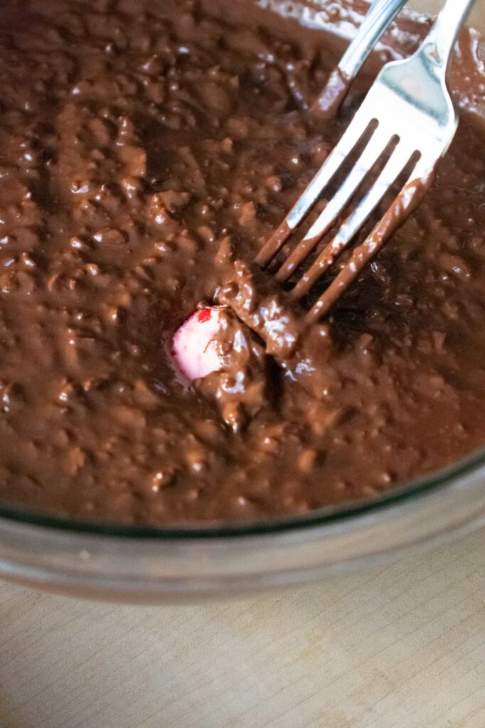 A fork dips a cherry mash candy center into textured chocolate in a glass bowl