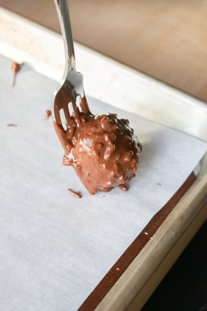 A fork holds a chocolate covered cherry mash candy over a parchment paper lined baking tray. Placing candy on tray to set.