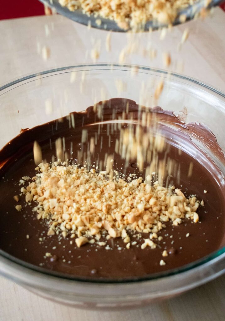Crushed nuts are sprinkled over a large glass bowl filled with smooth, melted chocolate while making candy coating for cherry mash candy recipe