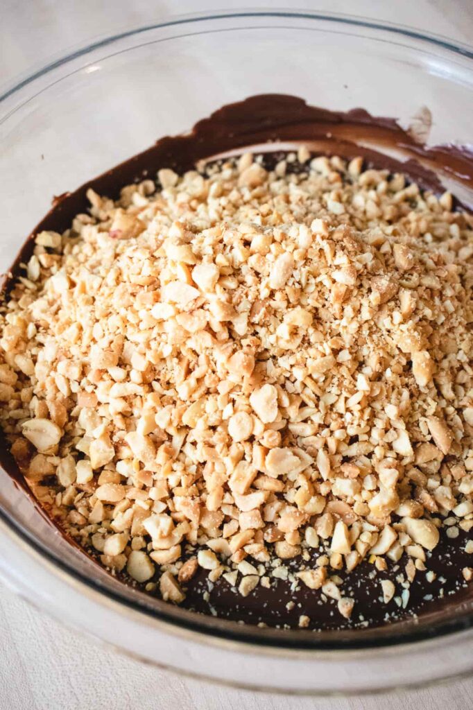 Close-up of a glass bowl filled with a rich, dark chocolate mixture topped with a generous layer of chopped, golden peanuts ready to mix.