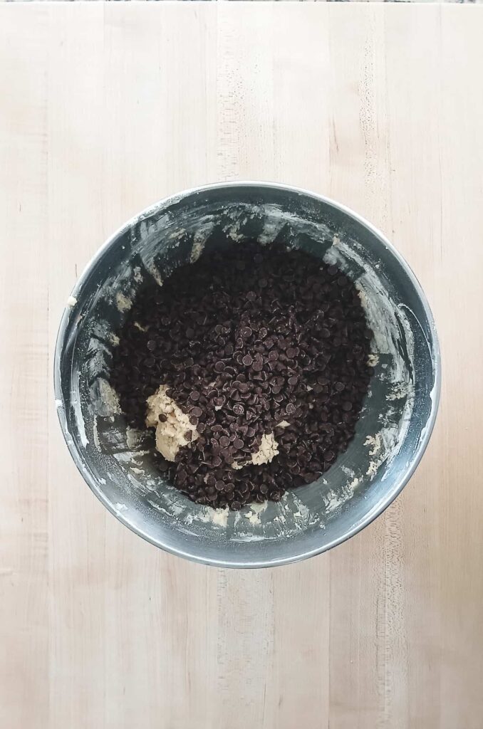 A large mixing bowl of pale cookie dough topped with a generous layer of mini semi-sweet chocolate chips before being folded together.